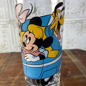May include: A clear glass drinking glass with a blue, yellow, black, and pink cartoon image of Mickey Mouse, Minnie Mouse, Pluto, and a pink bow. The characters are in a blue car.