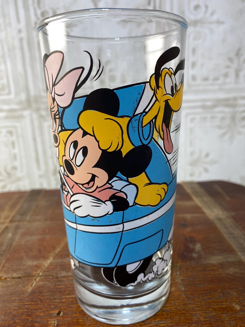 May include: A clear glass drinking glass with a blue, yellow, black, and pink cartoon image of Mickey Mouse, Minnie Mouse, Pluto, and a pink bow. The characters are in a blue car.