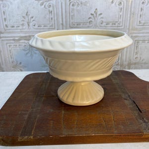 May include: Cream-colored pedestal bowl with a wide, shallow basin and a decorative, ridged base. The bowl features a textured, patterned design around the midsection. The bowl is made of ceramic and sits on a wooden surface.