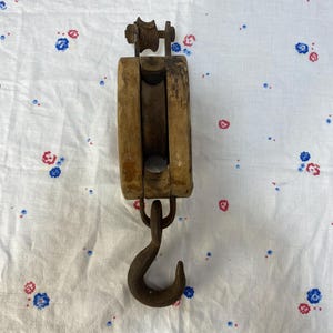May include: A vintage wooden pulley with a metal hook. The pulley has a rectangular wooden frame with a wheel at the top. The hook is curved and attached to the bottom of the frame. The pulley is likely used for lifting or pulling objects.