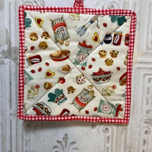 May include: Square quilted pot holder with a red and white gingham border. The cream-colored center features a repeating pattern of baking-themed illustrations, including strawberries, muffins, scales, and flour sacks.