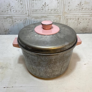 May include: Vintage metal cooking pot with a lid. The pot is silver with a textured finish. The lid has a pink handle and a pink rim. The handles on the side are also pink. The pot is sitting on a white surface.
