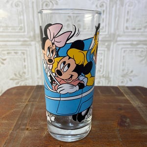 May include: A clear glass drinking glass with a blue, yellow, black, and pink cartoon image of Mickey Mouse, Minnie Mouse, and Goofy riding in a blue car.