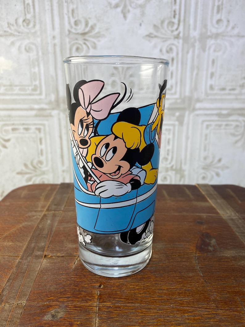 May include: A clear glass drinking glass with a blue, yellow, black, and pink cartoon image of Mickey Mouse, Minnie Mouse, and Goofy riding in a blue car.