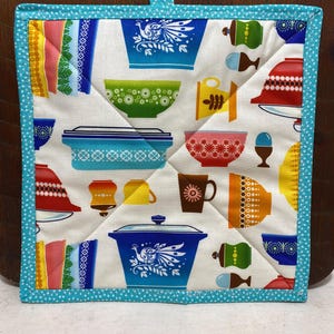 May include: A turquoise blue quilted hot pad with a white background and a pattern of colorful vintage kitchenware, including bowls, cups, and a casserole dish. The pattern features a peacock design on some of the dishes.