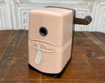 Puede incluir: Un sacapuntas vintage de color rosa con base negra y manivela de metal. El sacapuntas tiene las palabras "Boston Letter" impresas en el lateral.
