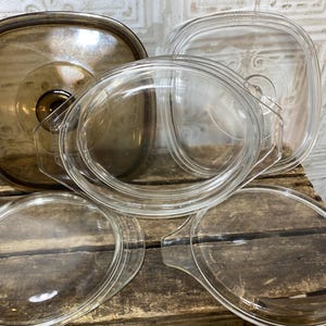 May include: A collection of four glass cookware lids, including one square amber-tinted lid and three clear glass lids in various shapes. The lids feature integrated handles and are arranged in a stack, with a yellow measuring tape visible.