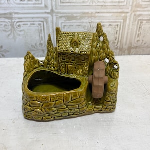 May include: A ceramic water mill planter in shades of green and brown. The planter features a small house, trees, and a water wheel. The base has a stone-like texture, and the interior is a darker green.