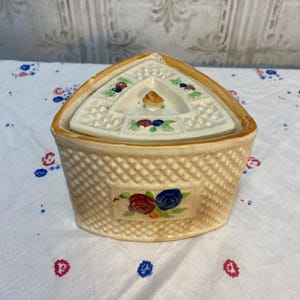 May include: A vintage, cream-colored ceramic basket-shaped container with a lid. The container has a woven texture and a rectangular floral design. The lid is triangular with a small handle and floral accents. The container sits on a white tablecloth with red and blue floral patterns.