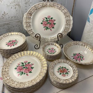 May include: A set of vintage white and gold floral dinnerware with pink roses. The set includes a large platter, a serving bowl, and several smaller plates.