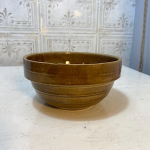 May include: A brown ceramic bowl with a glossy finish. The bowl has a wide, shallow shape with horizontal ridges around the exterior. The interior is a lighter shade of brown. The bowl is likely used for serving food or as a decorative piece.