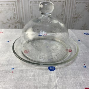 May include: A clear glass butter dish with a dome lid and a floral etched design. The dish is sitting on a white tablecloth with red and blue floral patterns.