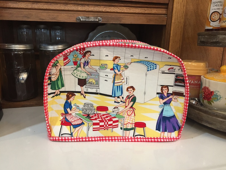 May include: A red and white checkered toaster cover with a vintage illustration of women baking in a kitchen. The cover features a yellow, white, and blue tiled floor, a red and white checkered tablecloth, and a red and white checkered border.
