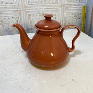 May include: A brown ceramic teapot with a matching lid. The teapot has a curved handle and a spout for pouring. The teapot is sitting on a white surface.