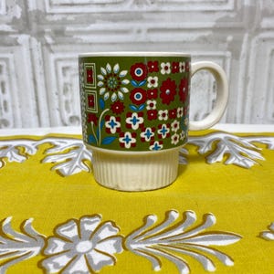 May include: A green ceramic mug with a multi-colored floral pattern. The mug has a white band around the middle and a white handle.