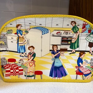 May include: A yellow and white toaster cover with a retro kitchen scene featuring women in vintage clothing. The scene includes a refrigerator, stove, and kitchen counter.