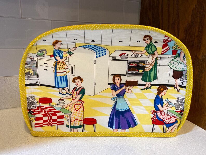 May include: A yellow and white toaster cover with a retro kitchen scene featuring women in vintage clothing. The scene includes a refrigerator, stove, and kitchen counter.