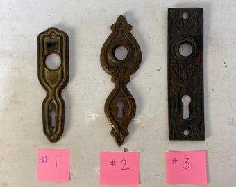 Puede incluir: Tres piezas antiguas de herrajes para puertas. Las piezas son de metal y tienen cerraduras y diseños decorativos. Cada pieza tiene una forma y un color diferentes, con una nota adhesiva rosa debajo con un número escrito.