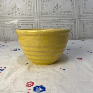 May include: A small, round, yellow bowl with horizontal ridges. The bowl has a slightly flared rim and sits on a white cloth with red and blue floral patterns. The background features a textured, off-white wall.