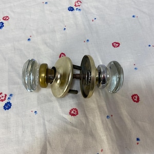 May include: A set of two glass door knobs with a brass and silver finish. The knobs are attached to a metal plate with screws.