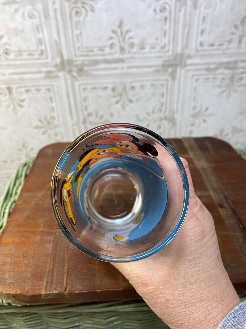 May include: A clear glass with a blue, yellow, and white design featuring Mickey Mouse and Minnie Mouse. The glass is upside down.