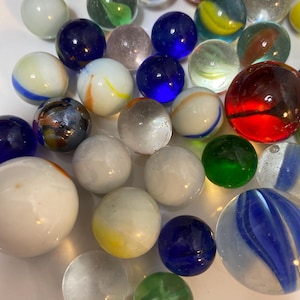 May include: A collection of glass marbles in various sizes and colors, including clear, solid white, deep blue, bright red, and green spheres. Some marbles feature internal swirls, streaks, or bubbles, while others are opaque or translucent glass.