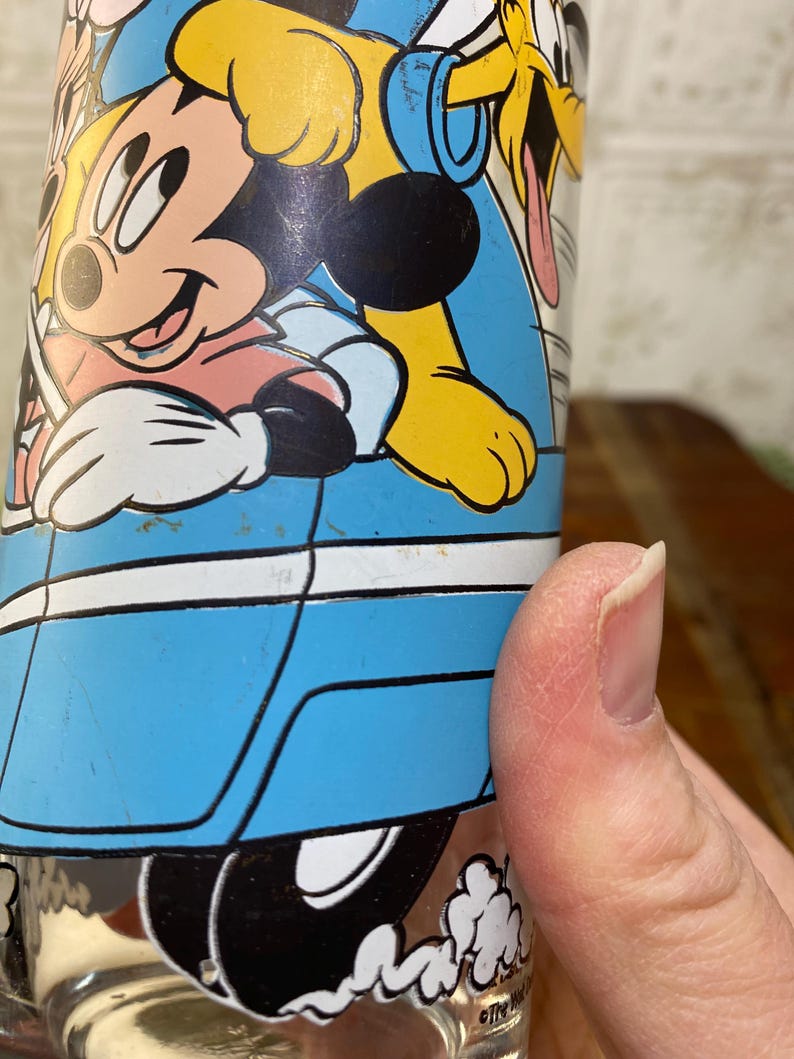 May include: A blue glass cup with a cartoon image of Mickey Mouse, Pluto, and Donald Duck. The characters are in a blue car with white trim. The car is driving on a white road with white clouds.