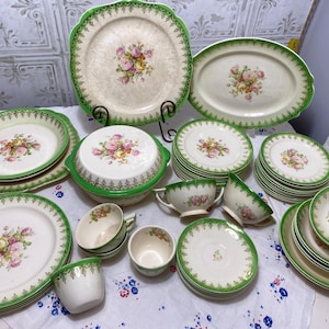 May include: A collection of vintage china with a floral rose pattern. The set includes plates, bowls, cups, and serving dishes with a cream background and green trim. The floral design features pink, yellow, and orange roses.