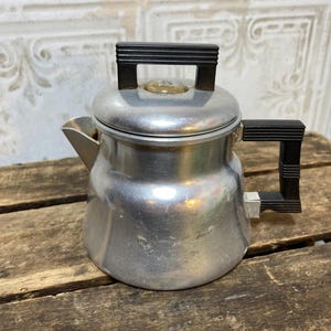 May include: A silver-colored aluminum stovetop coffee percolator featuring a rounded body, a short spout, and a removable lid. The percolator is equipped with a black, ribbed plastic handle on the side and a matching black, ribbed knob on the lid.