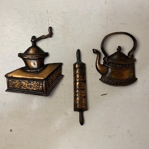 May include: Three antique bronze kitchen decor pieces: a coffee grinder, a rolling pin, and a teapot. The coffee grinder has a crank handle and sits on a rectangular base with floral detailing. The rolling pin is cylindrical. The teapot has a curved handle.