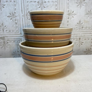 May include: A stack of three vintage mixing bowls. The bowls are cream-colored with horizontal stripes in shades of pink and blue. The bowls are nested inside each other, with the largest bowl at the bottom.