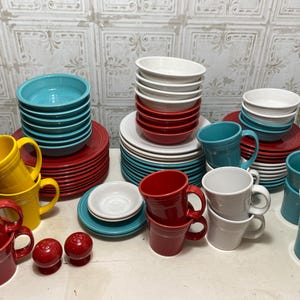 May include: A collection of colorful ceramic mugs and bowls in red, yellow, blue, and white. The mugs and bowls are stacked and arranged on a white surface.
