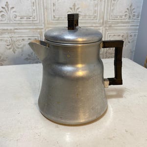 May include: A vintage aluminum coffee pot with a black handle and lid knob. The pot has a spout for pouring and a rounded body. The metal has a slightly worn appearance, suggesting age and use. The coffee pot is a classic kitchenware item.