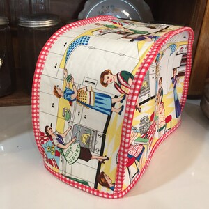 May include: A white toaster cover with a red and white checkered trim. The cover features a vintage illustration of women in a kitchen with a yellow and white checkered floor.