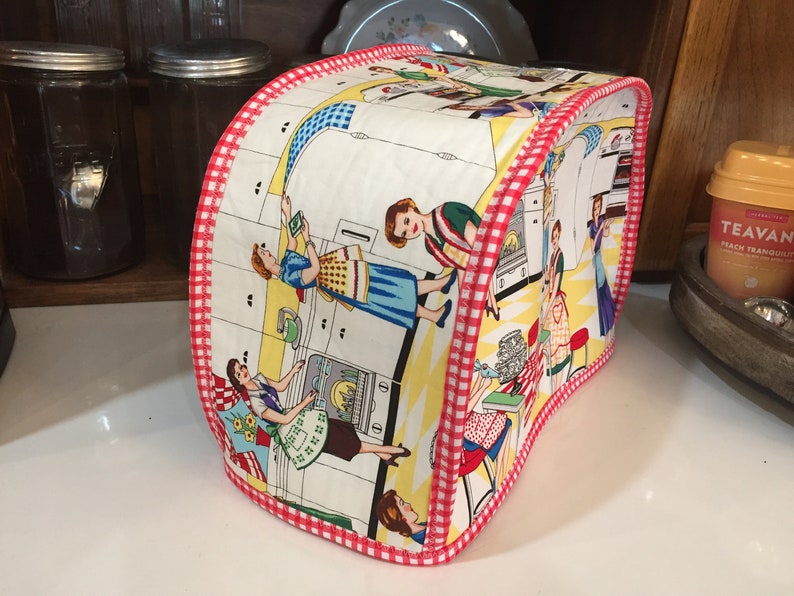 May include: A white toaster cover with a red and white checkered trim. The cover features a vintage illustration of women in a kitchen with a yellow and white checkered floor.