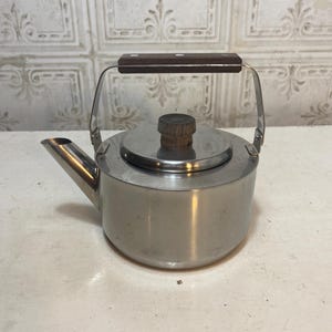 May include: A stainless steel tea kettle with a wooden handle and lid knob. The kettle has a spout for pouring and a handle that arches over the top. The background is a white surface with a decorative pattern.