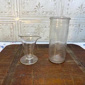 May include: Two clear glass measuring cups, one tall and one short, with graduated markings. The cups are sitting on a wooden surface.