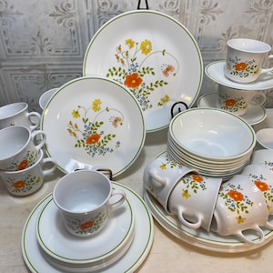 May include: A vintage white dinnerware set with a floral design. The set includes plates, bowls, and cups with a green trim. The floral pattern features orange, yellow, and blue flowers. The set is arranged on a white surface.