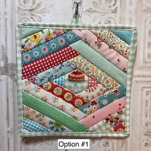 May include: A square quilted potholder featuring a diagonal strip pattern of various floral, gingham, and polka dot fabrics in shades of pink, green, blue, and red. The center displays a small illustration of a cake on a stand. Text reads Option #1.