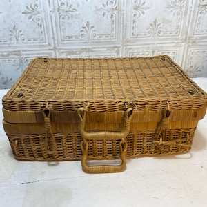 May include: A rectangular, light brown wicker picnic basket with a woven design. The basket has a hinged lid, metal clasps, and a woven handle. The basket is sitting on a white surface.