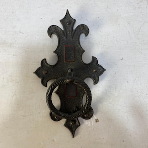 May include: An antique-style door knocker made of dark metal, featuring a decorative backplate and a twisted ring handle. The design is symmetrical with pointed details and small brass studs. The backplate has a red square in the center.