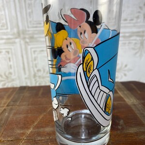 May include: A clear glass drinking glass with a cartoon image of Mickey Mouse, Minnie Mouse, and Donald Duck in a blue car. The car is driving fast and there are white clouds behind it.