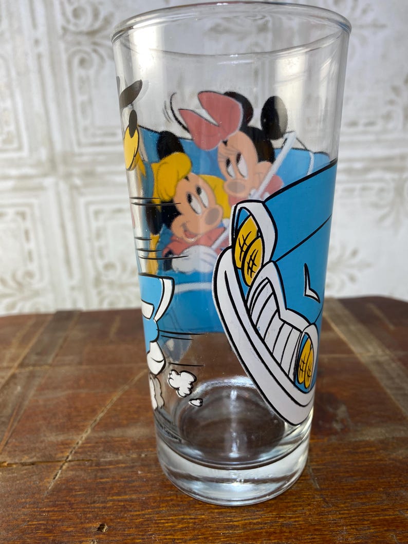 May include: A clear glass drinking glass with a cartoon image of Mickey Mouse, Minnie Mouse, and Donald Duck in a blue car. The car is driving fast and there are white clouds behind it.