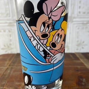 May include: A vintage glass drinking glass with a blue car design featuring Mickey Mouse and Minnie Mouse. The car has white wheels and a white exhaust trail. The glass has a white rim.