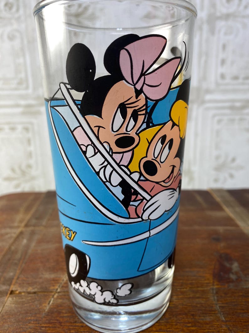 May include: A vintage glass drinking glass with a blue car design featuring Mickey Mouse and Minnie Mouse. The car has white wheels and a white exhaust trail. The glass has a white rim.