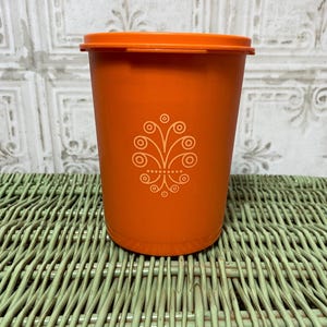 May include: An orange plastic container with a matching lid. The container has a white floral design on the front. The container is sitting on a green woven surface.