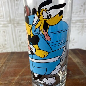 May include: A vintage glass drinking glass with a blue, yellow, and black cartoon image of Mickey Mouse and Pluto driving a blue car. The text "The Walt Disney Co." is printed on the bottom of the glass.