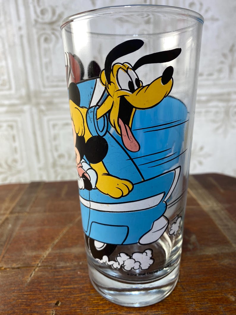 May include: A vintage glass drinking glass with a blue, yellow, and black cartoon image of Mickey Mouse and Pluto driving a blue car. The text "The Walt Disney Co." is printed on the bottom of the glass.