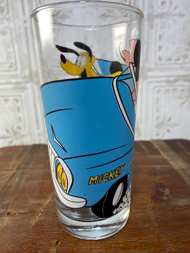 May include: A blue glass with a cartoon image of Mickey Mouse, Pluto, and Goofy driving a blue car. The car has the word "MICKEY" printed on the side.