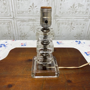 May include: A clear glass table lamp with a square base and stacked, rectangular blocks. The lamp has a silver-colored metal socket and a brown, cylindrical top. A cream-colored cord extends from the base. The lamp is on a wooden surface.